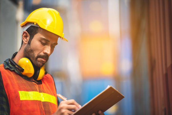 A construction worker in an orange vest and hard hat takes notes on a clipboard while standing outdoors.