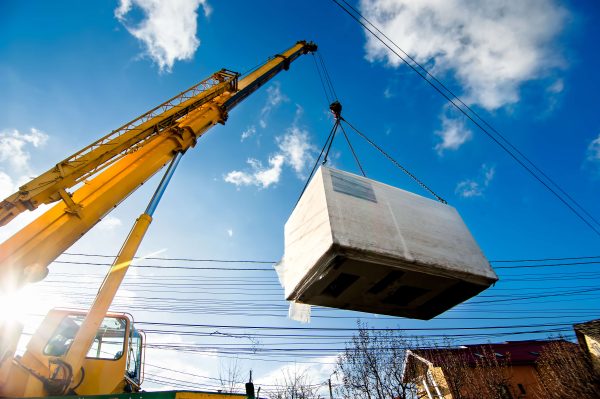 A crane lifts a large white box onto a truck at a construction site, showcasing heavy machinery in action.